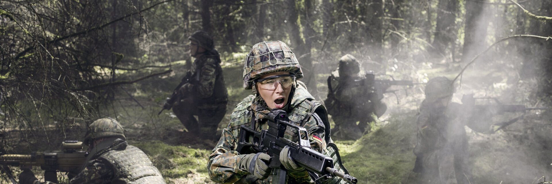 Soldatinnen und Soldaten liegen bei einer Übung auf dem Waldboden und sichern mit Gewehren die Umgebung.